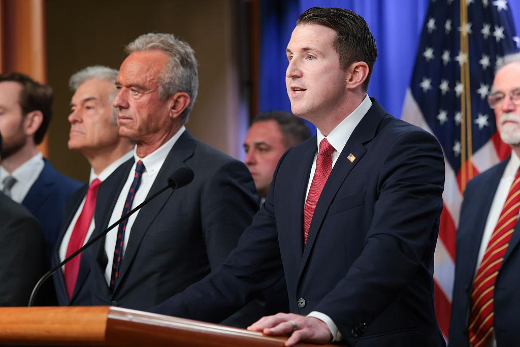 Officials at a press conference, with a speaker at a podium and US flags in the background.